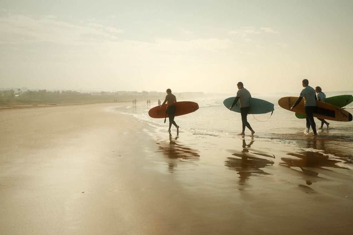 Mehrere Surfer tragen ihre Bretter bei tief stehender Sonne über den nassen Strand.