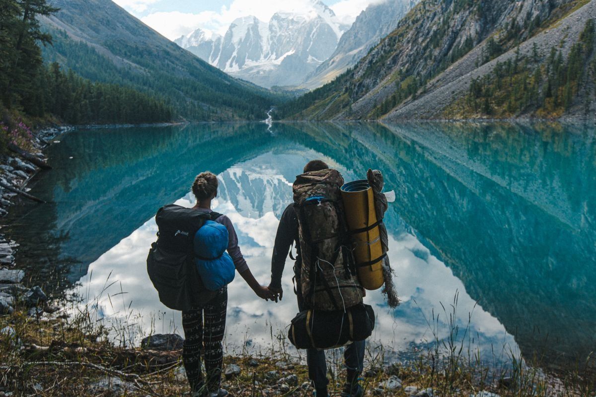 Paar mit großen Rucksäcken steht Hand in Hand an einem türkisblauen Bergsee