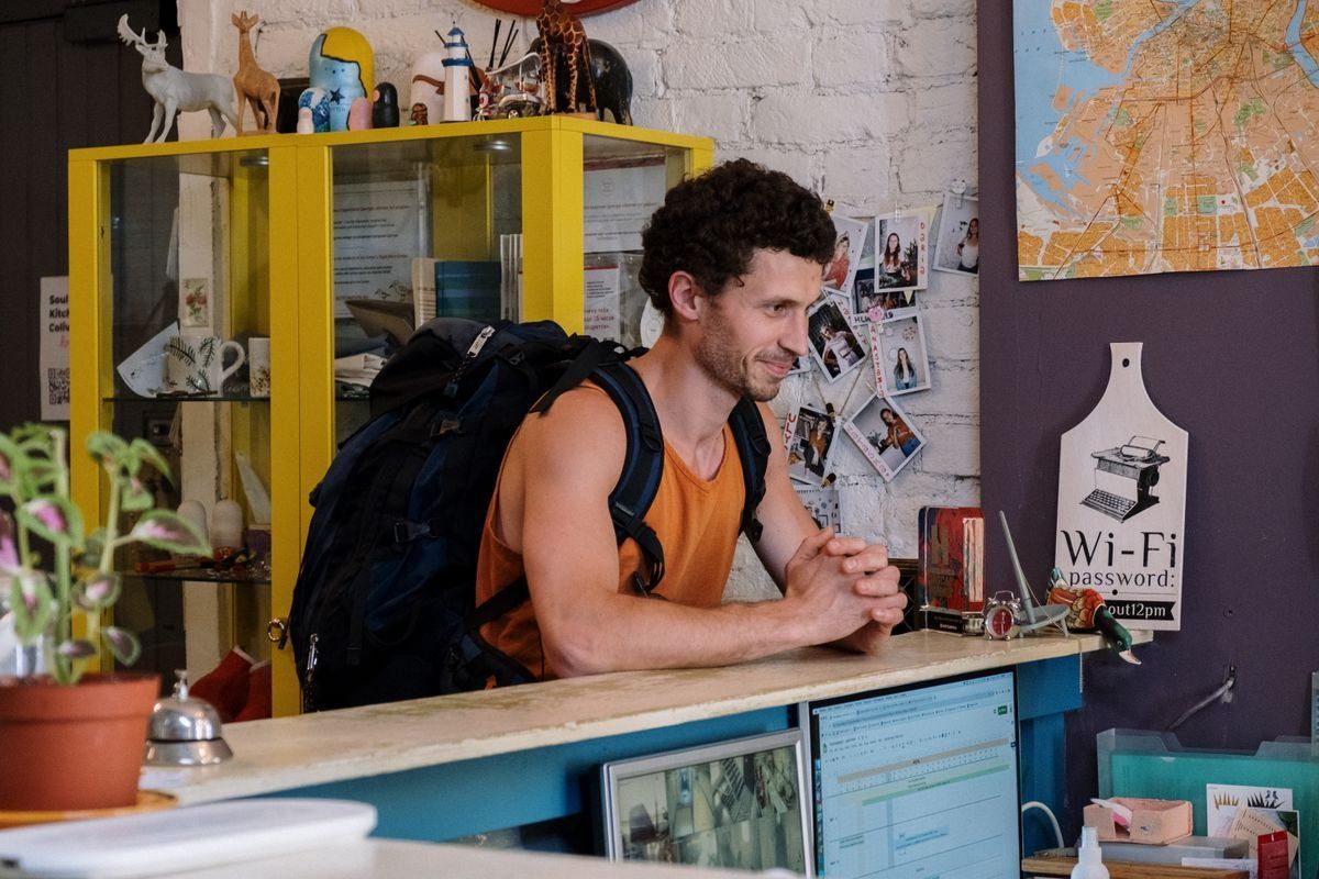 Reisender mit großem Rucksack an einer Hostel-Rezeption mit bunten Regalen im Hintergrund