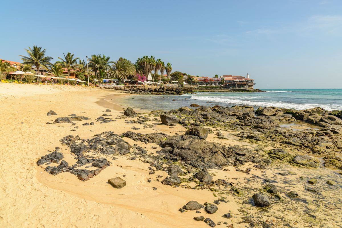 Sandstrand mit schwarzen Felsen, Palmen und kleinen Wellen unter klarem blauem Himmel
