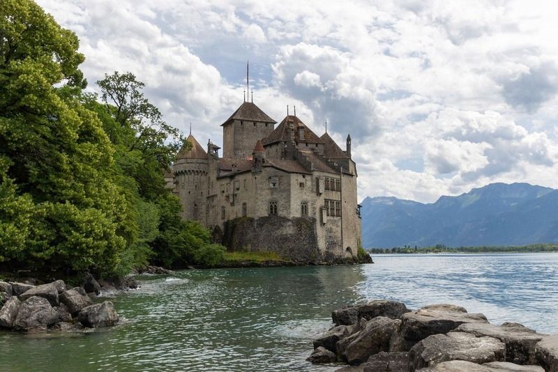 Schloss Chillon am Ufer des Genfersees, umgeben von Felsen und Grün, mit den Alpen im Hintergrund