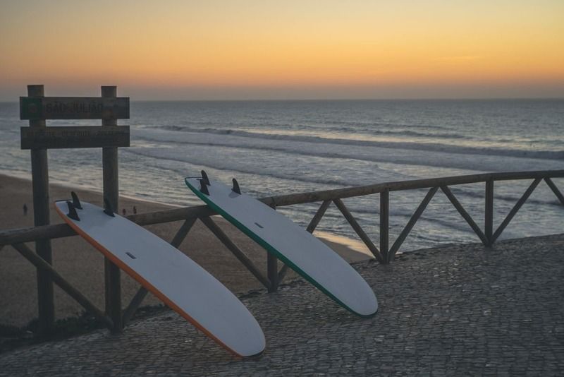 Zwei Surfbretter lehnen an einem Holzgeländer vor dem Meer, Schild „São Julião" im Sonnenuntergang