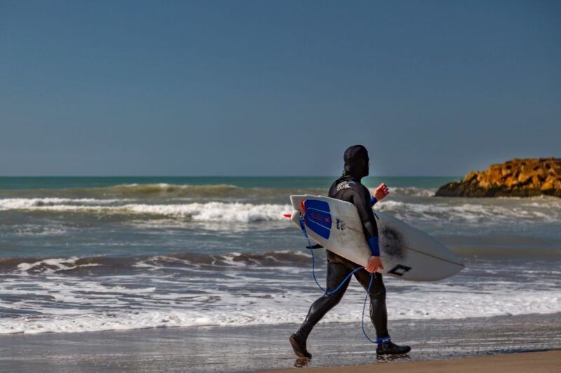 Surfer im Neoprenanzug läuft mit Surfbrett am nassen Strand entlang.
