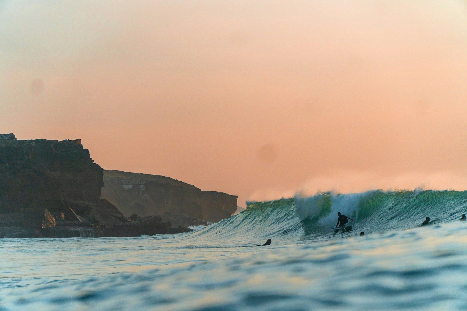 Surfer auf einer Welle bei Sonnenuntergang vor der Silhouette der Steilküste Portugals
