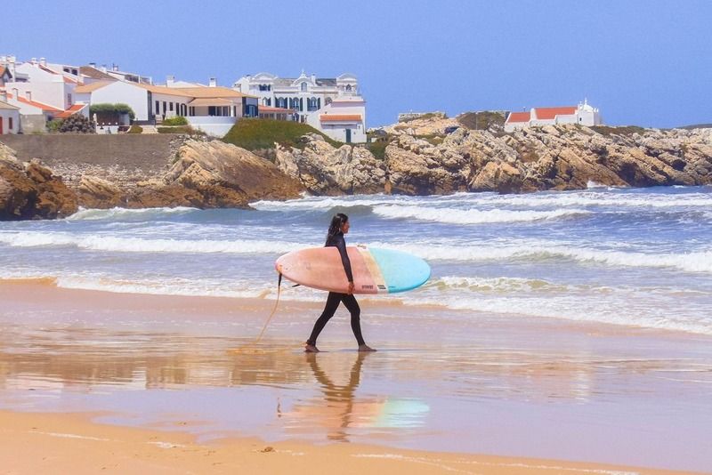 Surferin mit Surfbrett am Strand, im Hintergrund ein weißes portugiesisches Dorf auf der Klippe