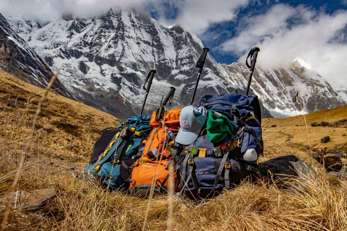 Mehrere Trekkingrucksäcke und Stöcke auf trockenem Gras vor schneebedeckten Bergen