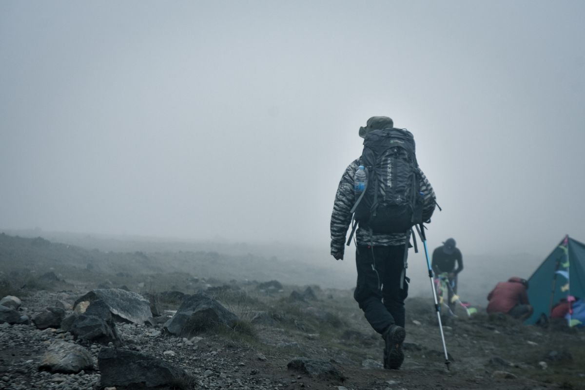 Wanderer mit großem Rucksack und Stock in nebliger felsiger Berglandschaft neben Zelten