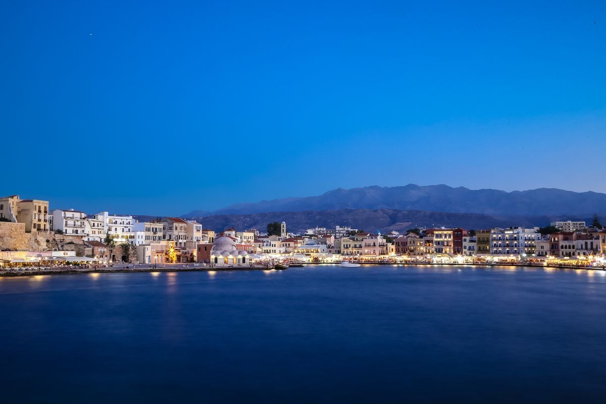 Hafen von Chania in der Abenddämmerung mit beleuchteten Gebäuden und Bergen am Horizont