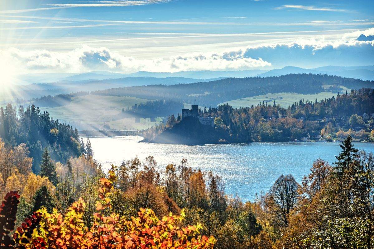 Sonnenstrahlen über einem See mit herbstlichen Wäldern und einer Burg auf einem Hügel