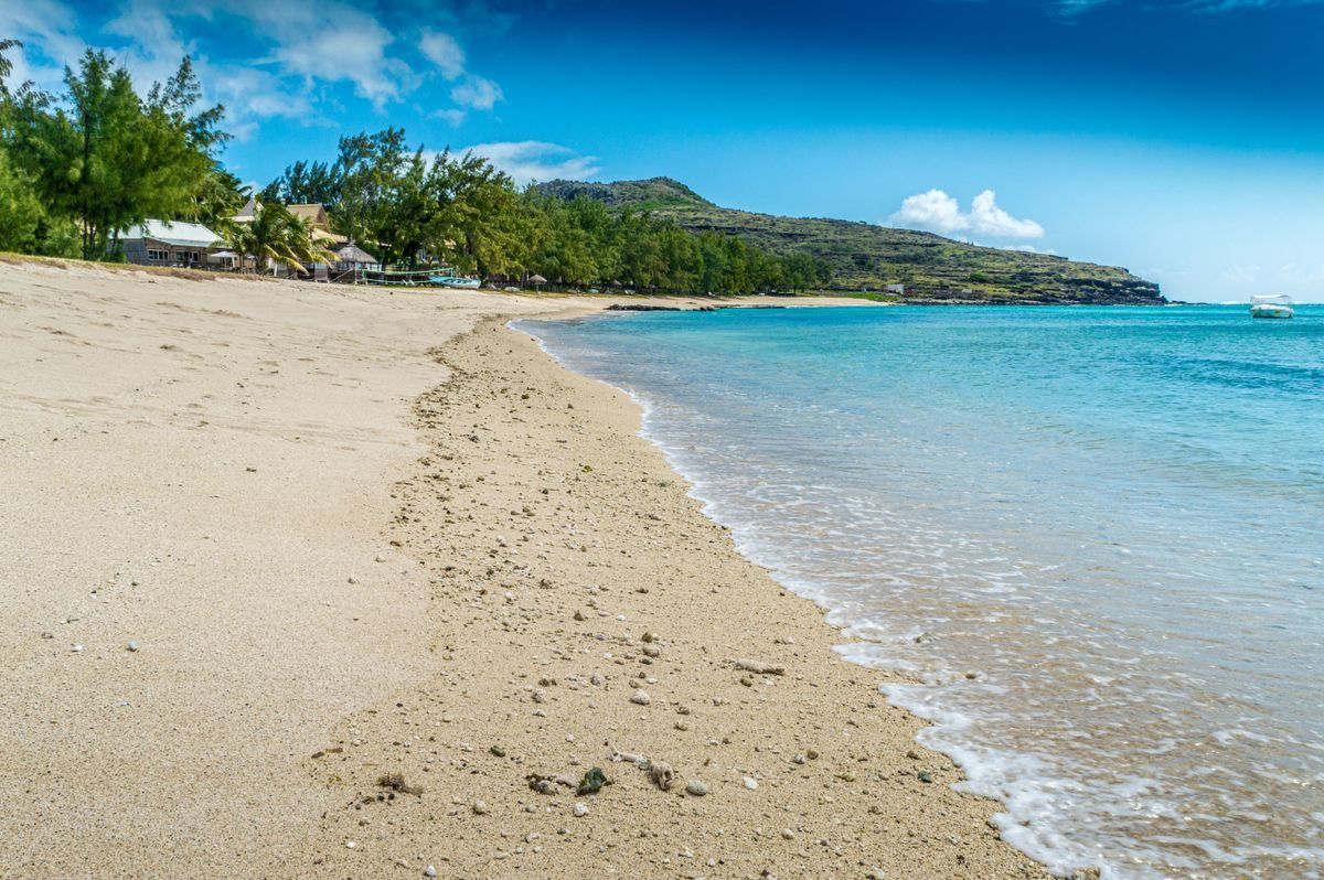 Breiter Sandstrand mit flachem klarem Wasser, Bäumen und grünem Hügel im Hintergrund