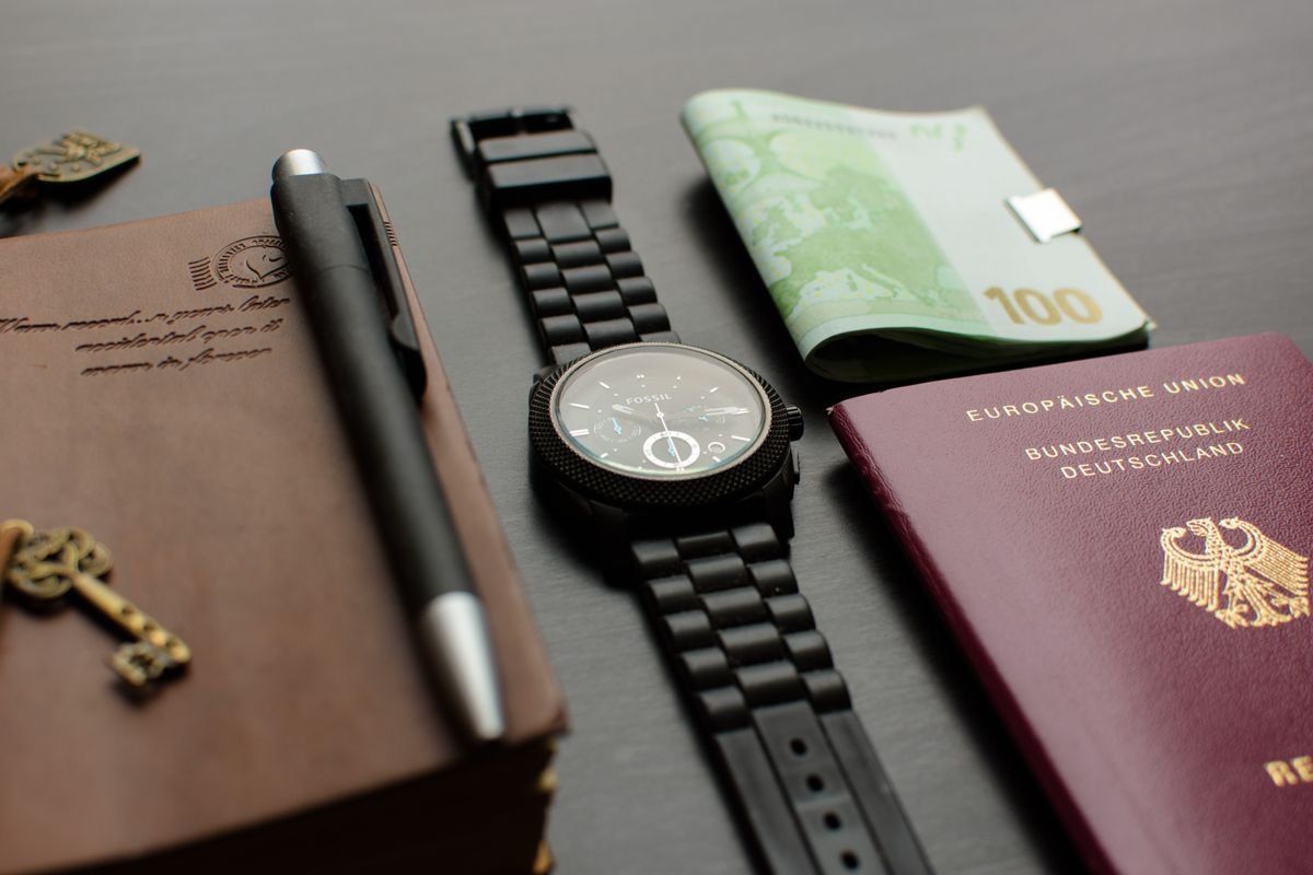 Deutscher Reisepass mit Armbanduhr, Geldschein, Notizbuch und Stift auf dunklem Tisch