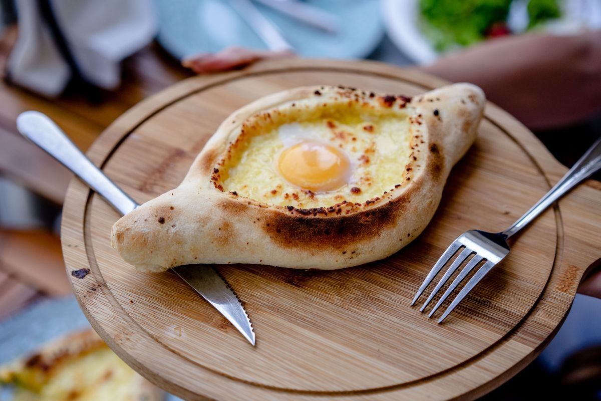 Khachapuri mit Ei auf runder Holzplatte, daneben Messer und Gabel